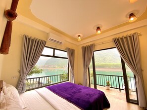 A hotel room with a bed, large windows, and a balcony with a view of the water and mountains. - Phong Nha Coco Riverside Homestay (Bo Trach)