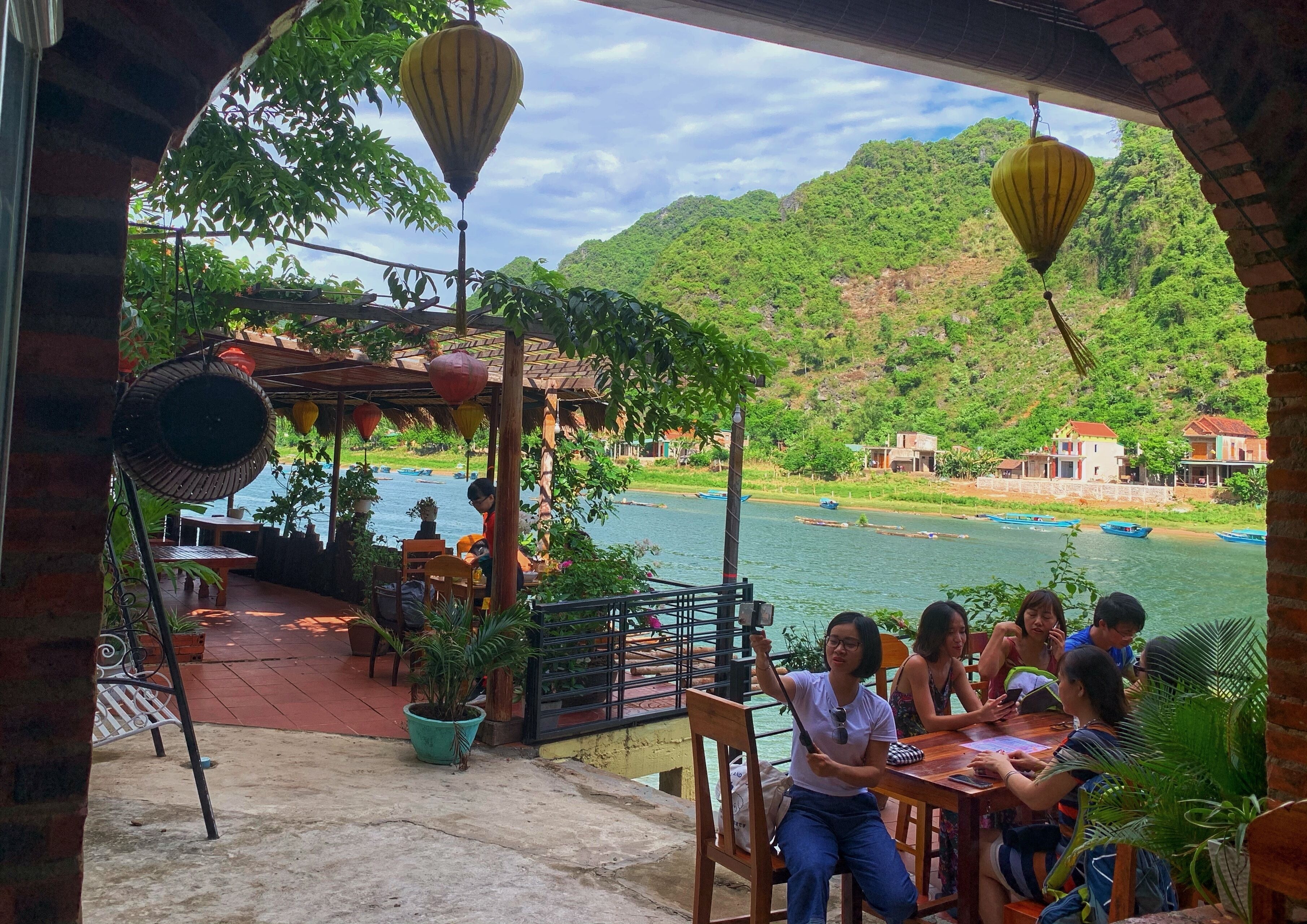 A riverside restaurant with outdoor seating, overlooking a river and hills.