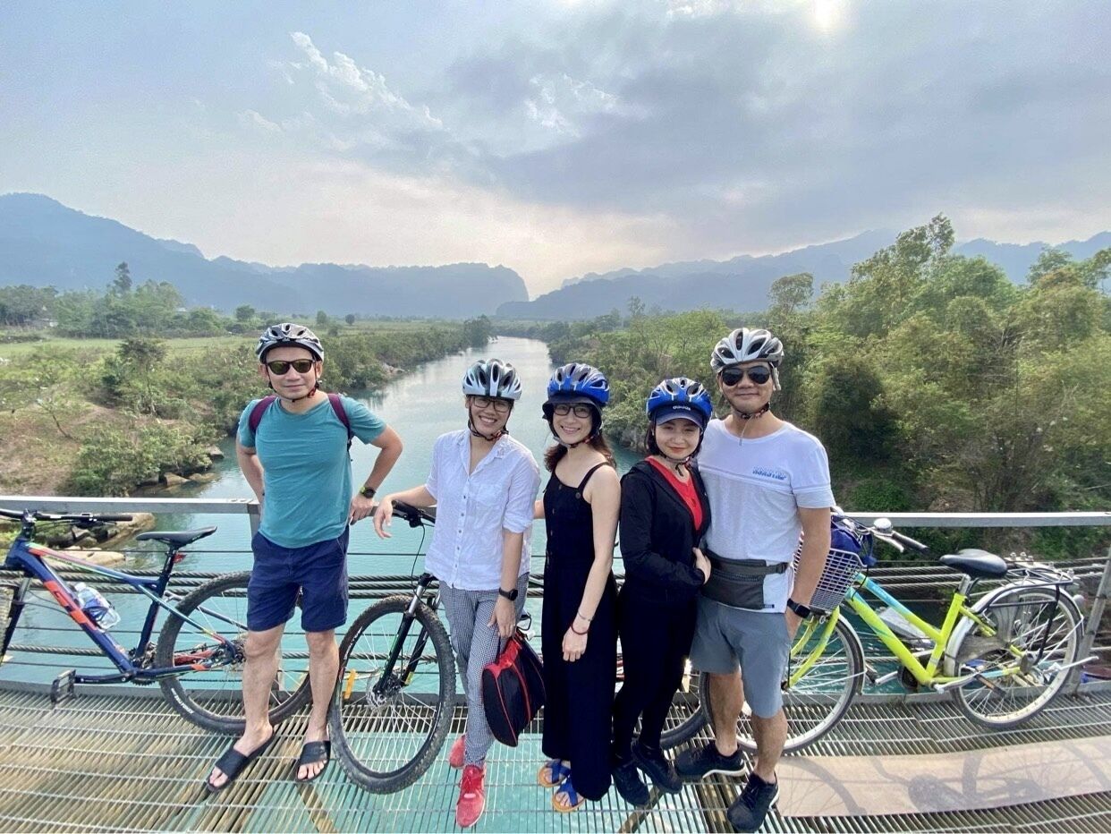 A group of people on a bridge with bicycles, a river, and mountains in the background.