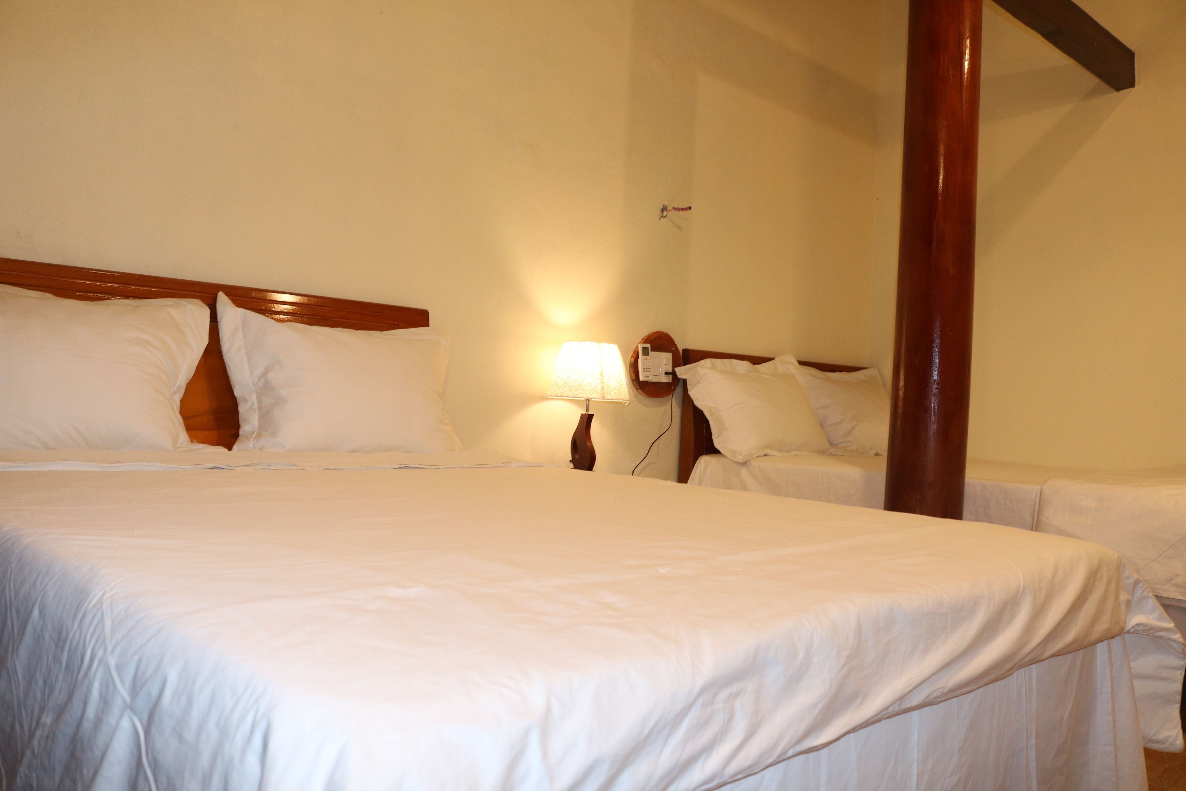 Een hotelkamer met een groot bed, twee kleinere bedden, een houten hoofdeinde, een nachtkastje met een lamp en een houten zuil.