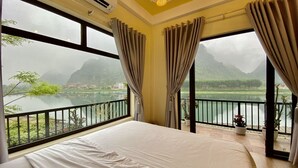 A bedroom with a large bed, a balcony with a view of a lake and mountains, and an air conditioning unit. - Phong Nha Coco Riverside Homestay (Phong Nha)