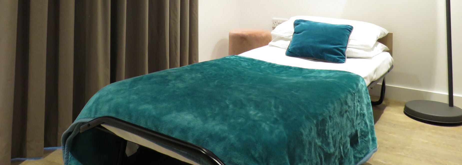 Una habitació d'hotel amb un llit, una manta color turquesa, un coixí, una taula de nit i una làmpada de peu.
