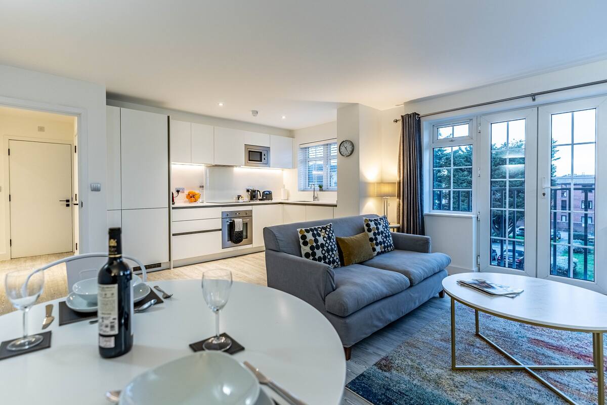 A modern living room with a grey sofa, a round dining table with wine glasses and a bottle, and a view through large windows.