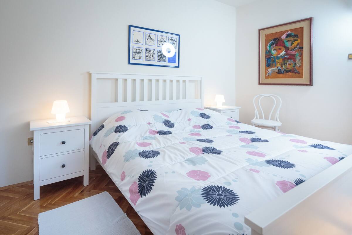 Ein Schlafzimmer mit einem weißen Bett, zwei Nachttischen, einem weißen Stuhl, einem gerahmten Bild an der Wand und einem farbenfrohen abstrakten Gemälde.