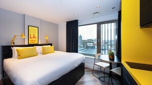 Una habitación de hotel moderna con una cama grande, una pequeña mesa de comedor, una silla y vistas a la ciudad a través de amplios ventanales.
