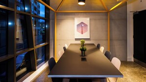 Un salón de conferencias con una larga mesa, sillas, una planta en maceta, una obra de arte enmarcada y amplias ventanas.