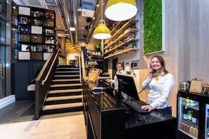 Zona de recepción de un hotel con escalera, mostrador de recepción, computadora y refrigerador con bebidas.