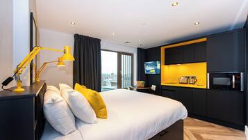 Una habitación de hotel moderna con una cama grande, un escritorio con lámpara, una kitchenette con microondas y un televisor en la pared.