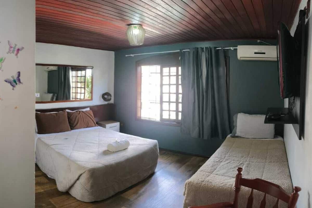 A bedroom with a bed, a bedside table, a mirror, a window with curtains, and a wooden ceiling.