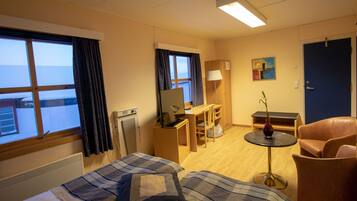 A hotel room with a bed, a desk, a chair, a small table with a vase, and a view of the sea through the window.