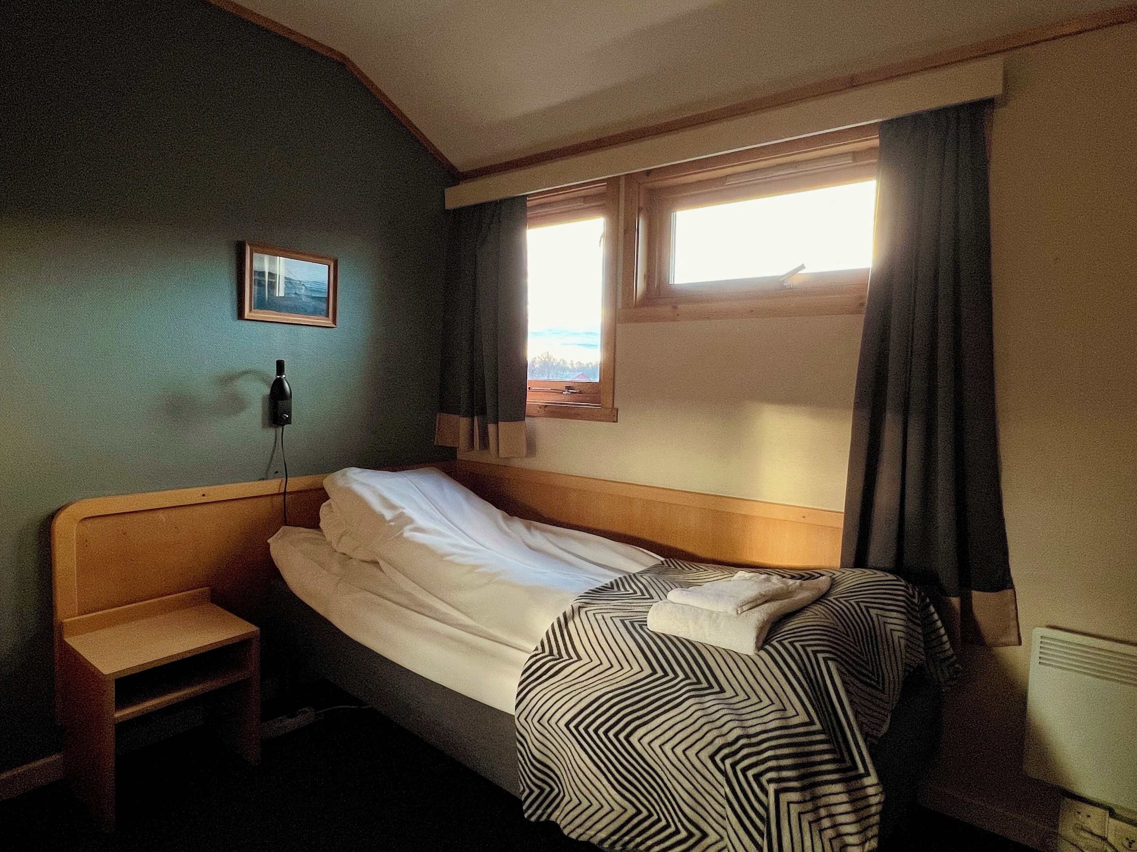 A single bed with a striped blanket, a bedside table, a wall-mounted phone, and a window with curtains.