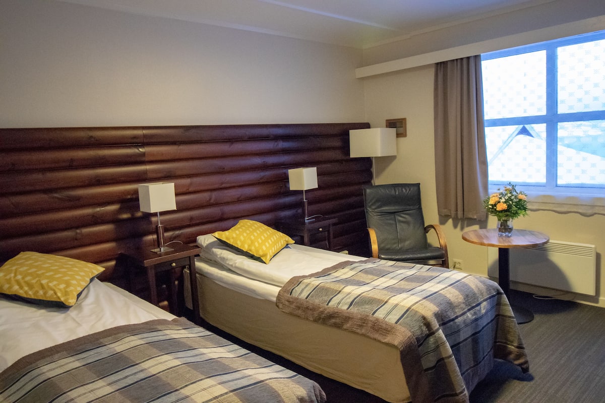 A hotel room with two beds, a wooden headboard, a small table with a vase, and a window with curtains.