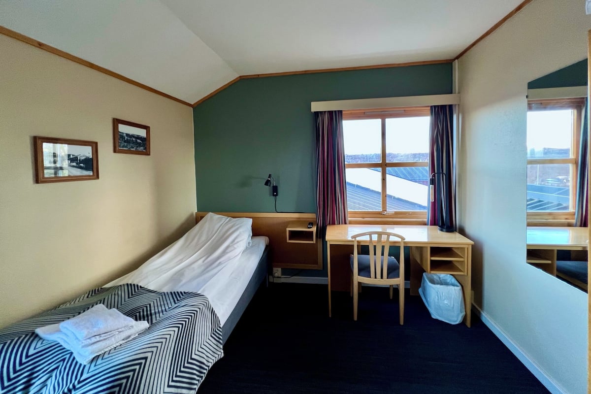 A small, neatly arranged room with a bed, a desk, and a chair. There are two framed pictures on the wall, a window with curtains, and a view of a snowy landscape outside.