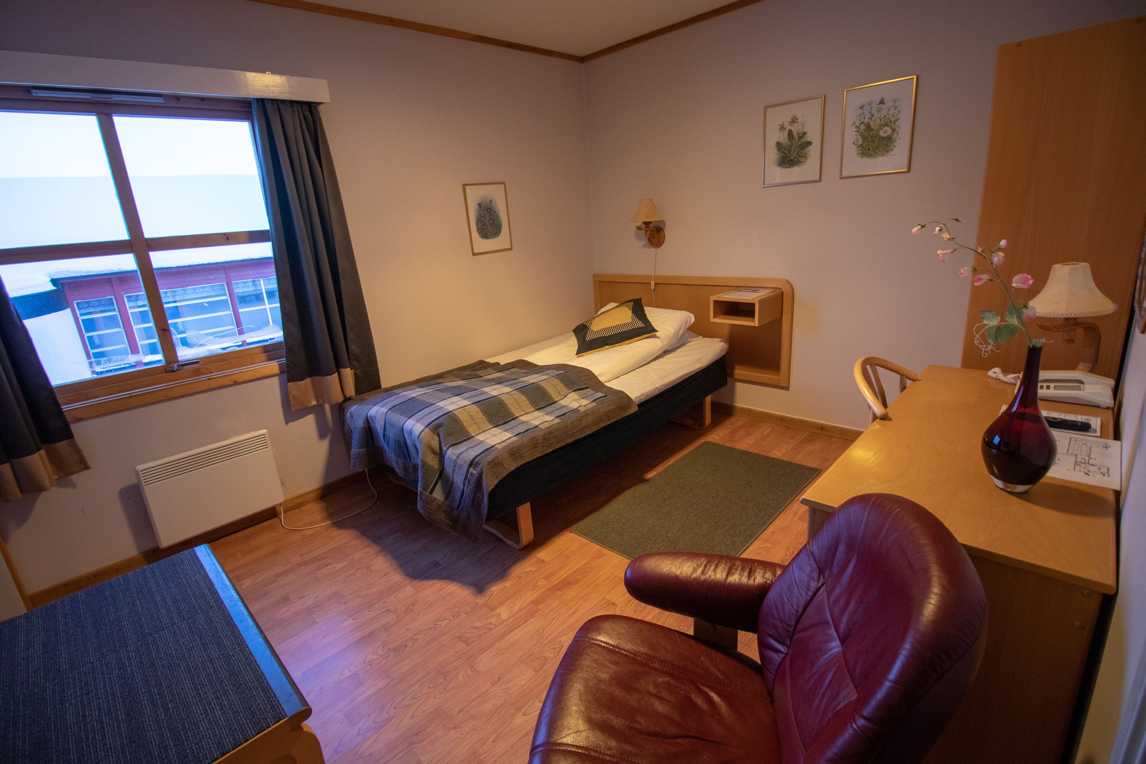 A hotel room with a bed, a desk, a chair, a window with curtains, a radiator, and a wall-mounted lamp.