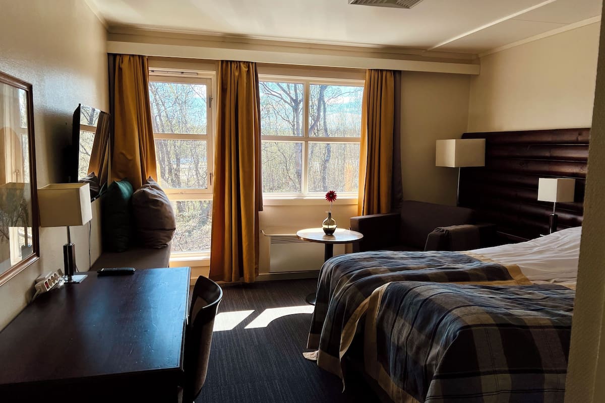 A hotel room with a bed, a desk, a chair, a small table with a vase, and a window with curtains.