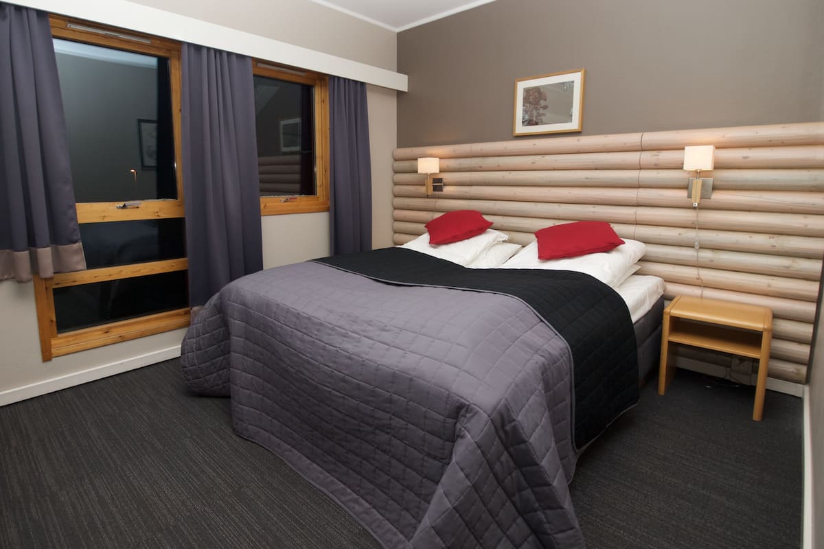 A hotel room with a bed, a window with curtains, a bedside table, and a wall-mounted lamp.