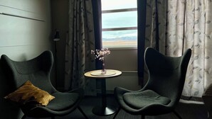 Et værelse med to mørkegrønne lænestole, et rundt bord med en blomstervase, og udsigt over et landskab gennem et vindue med mønstrede gardiner.