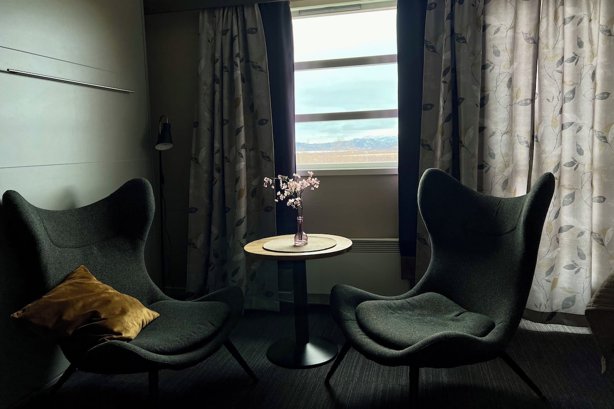 A room with two dark green armchairs, a small round table with a vase of flowers, and a view of a landscape through a window with patterned curtains.