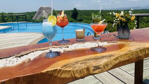 Una barra de madera con vista a la piscina, mostrando cuatro bebidas y un jarrón con flores.