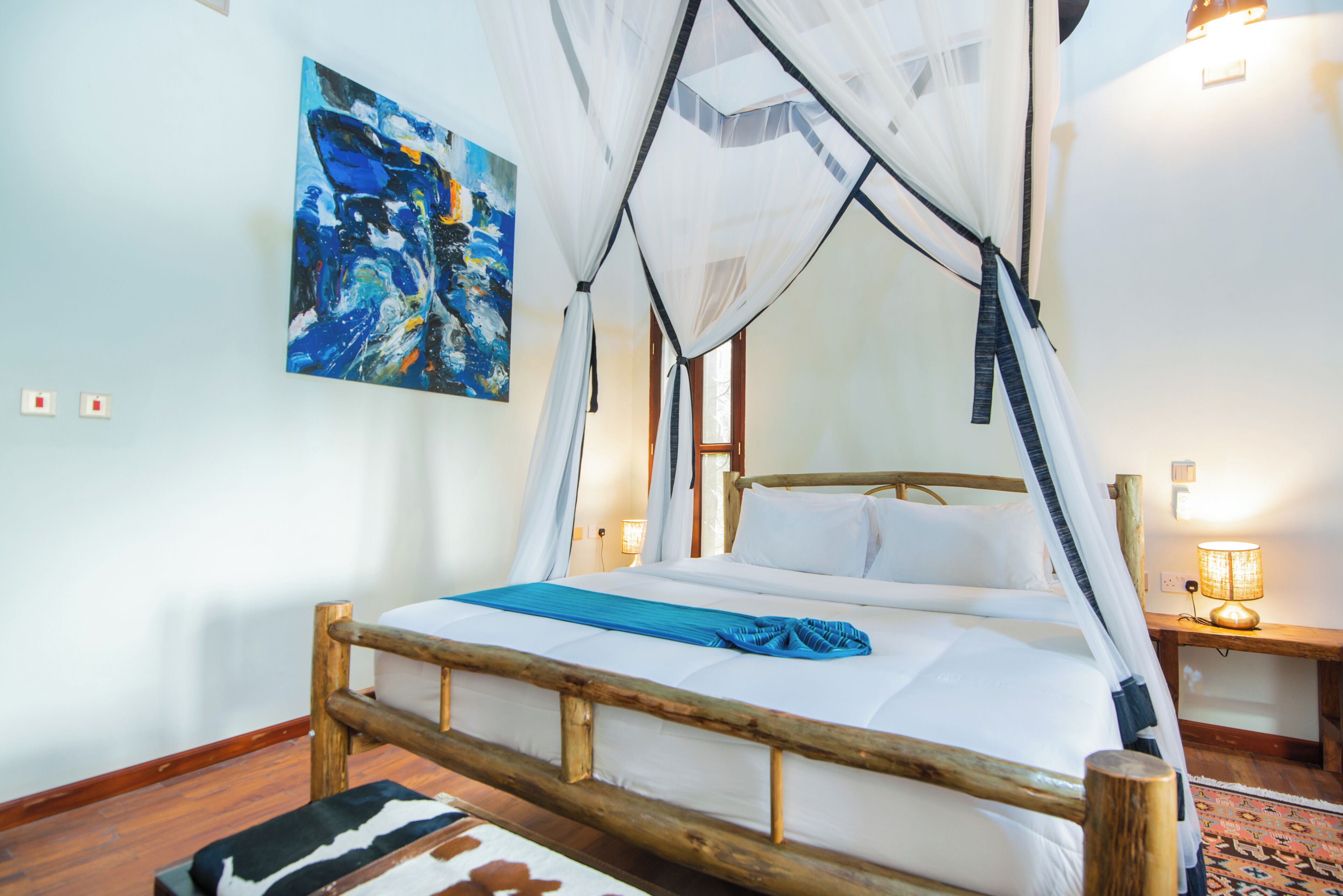 Un lit avec un cadre en bois, une literie blanche et un plaid bleu. Il y a un tableau au mur et une lampe sur une table de chevet.