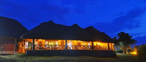 A thatched-roof building with a wooden fence and a stone wall, illuminated by warm lights at night.