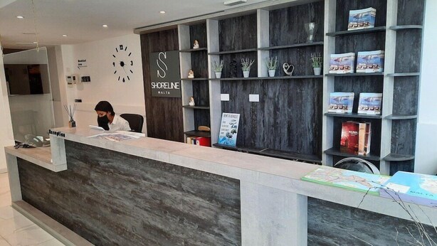 A reception desk with a modern design, featuring a dark wood panel and a sign that reads "Shoreline Malta". There are shelves with books and decorative items, and a clock on the wall. - Gold Lion Residensea (St. Julian's)