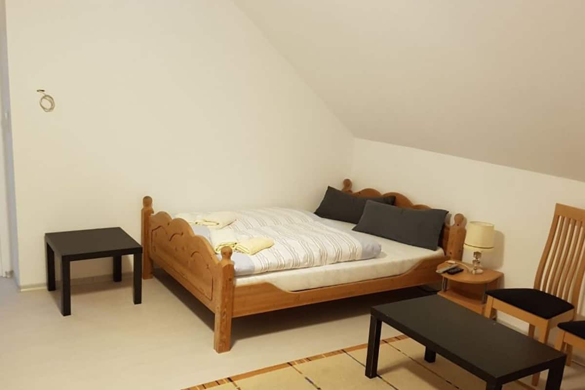 A bedroom with a wooden bed, a bedside table, a chair, a small table, and a lamp.