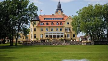 Stately building with a prominent tower and central entrance, set amidst a tranquil lawn with a fountain.