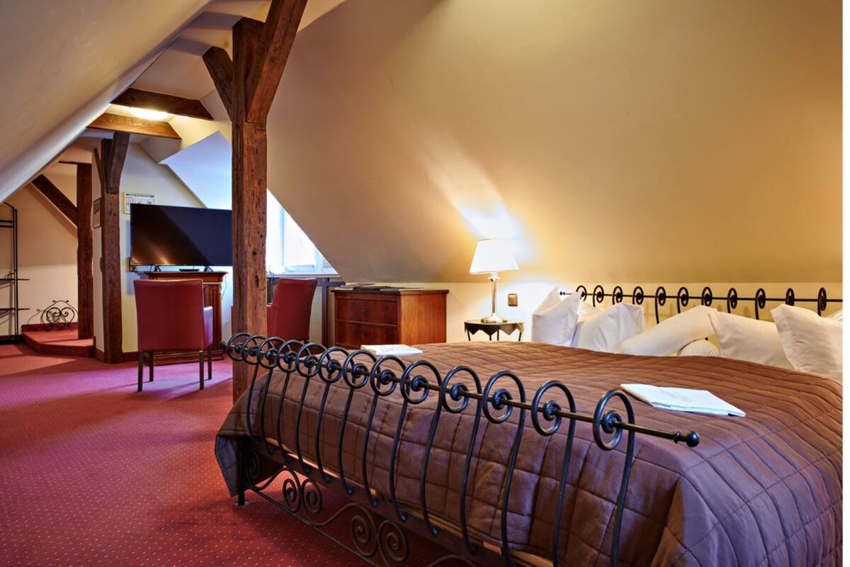 Une chambre avec un plafond en pente, un lit avec un cadre en métal, une chaise en bois, un bureau et une lampe.