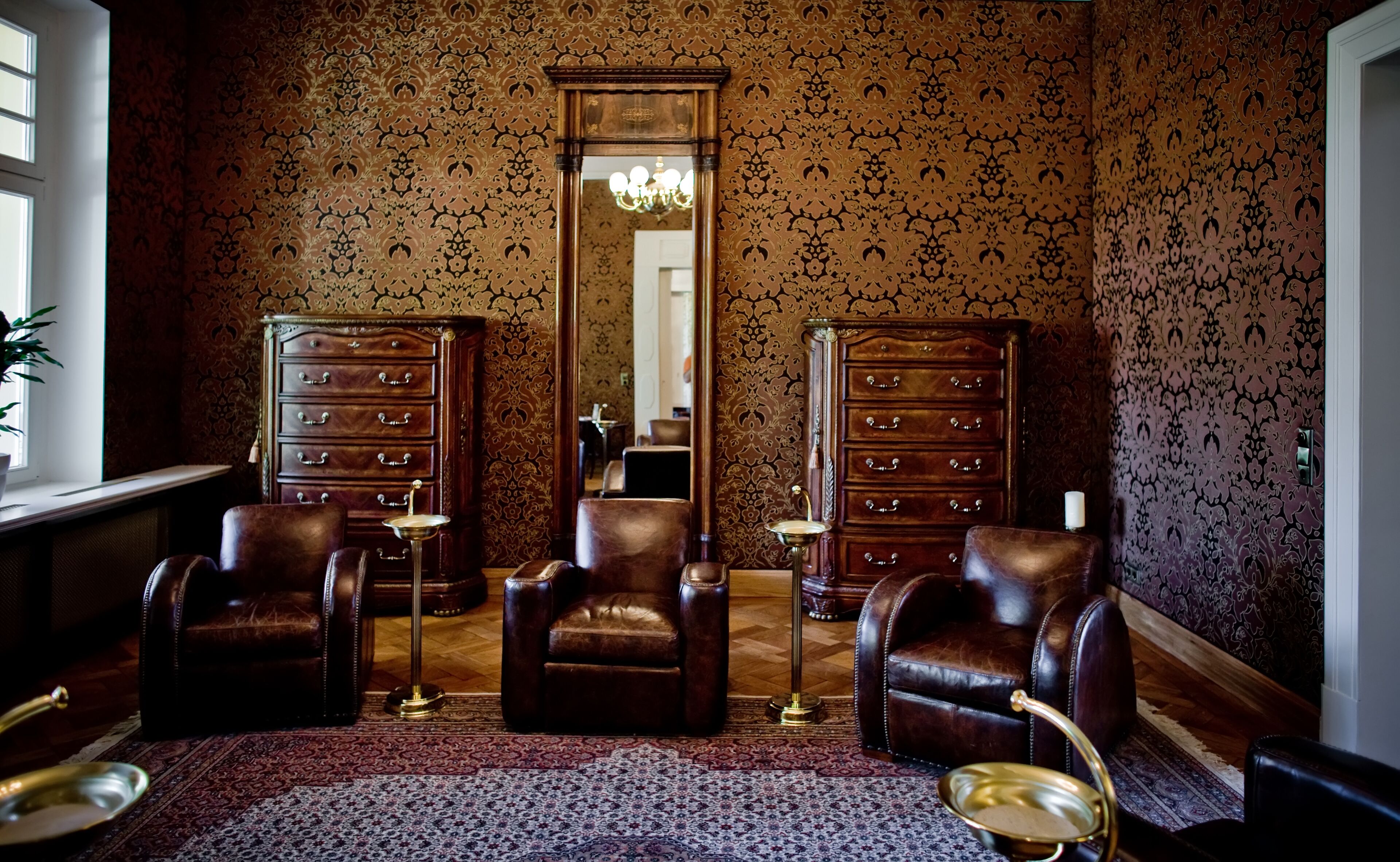 A room with a patterned wallpaper, two wooden dressers, leather chairs, a chandelier, and a large mirror.