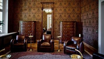 A room with a patterned wallpaper, two wooden dressers, leather chairs, a chandelier, and a large mirror.
