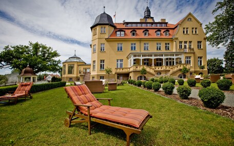 A large, yellow building with a red-tiled roof, a tower, and a well-maintained lawn with lounge chairs.
