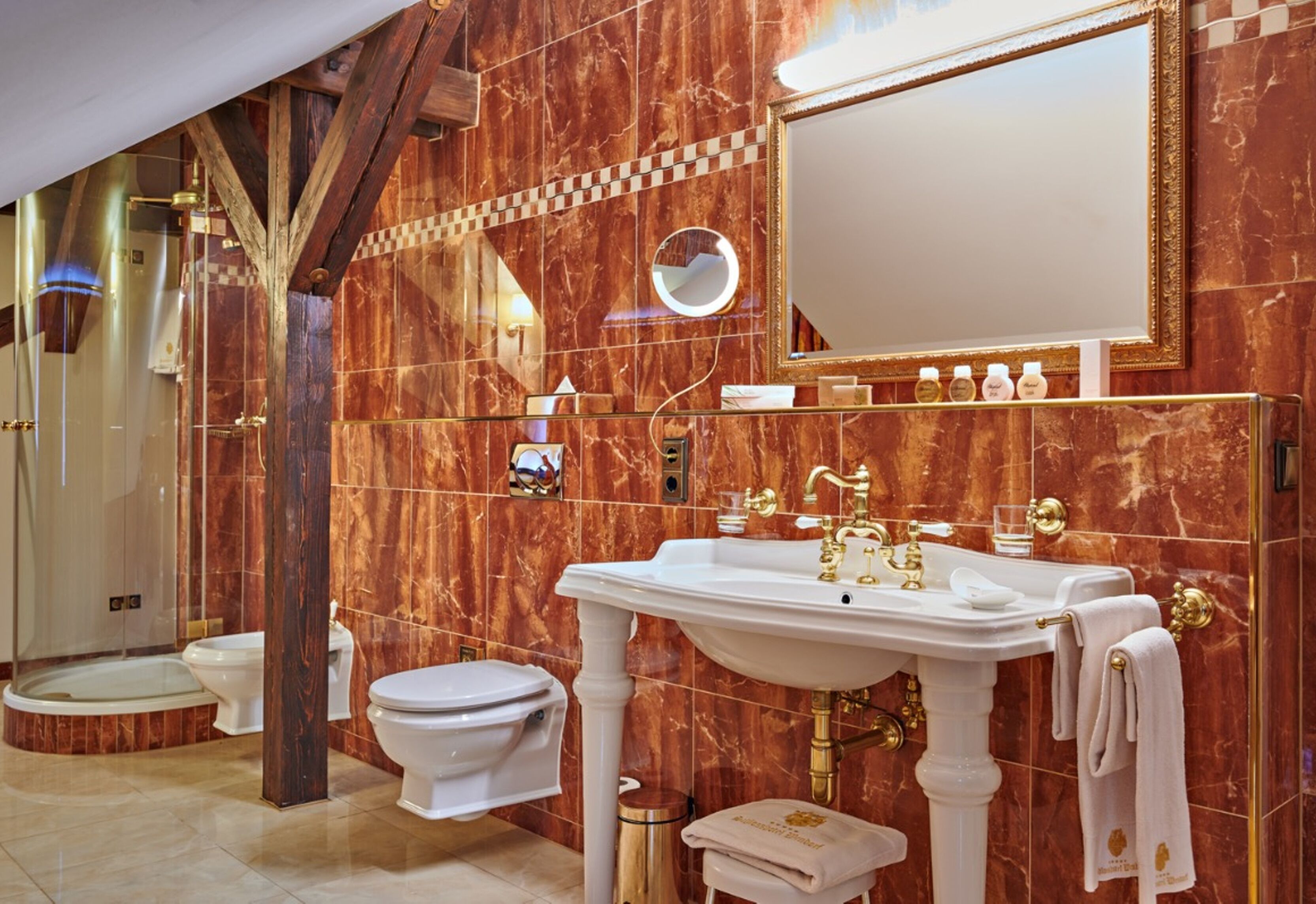 Un baño lujoso con pared de mármol, un espejo grande, un lavabo blanco con detalles dorados y un inodoro.