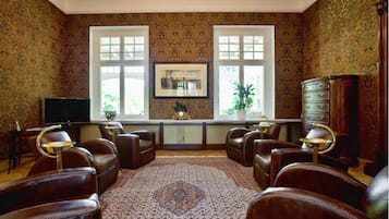 Une pièce dotée d’un tapis à motifs, de fauteuils en cuir, d’un téléviseur, d’une armoire et de deux fenêtres ornées de plantes.