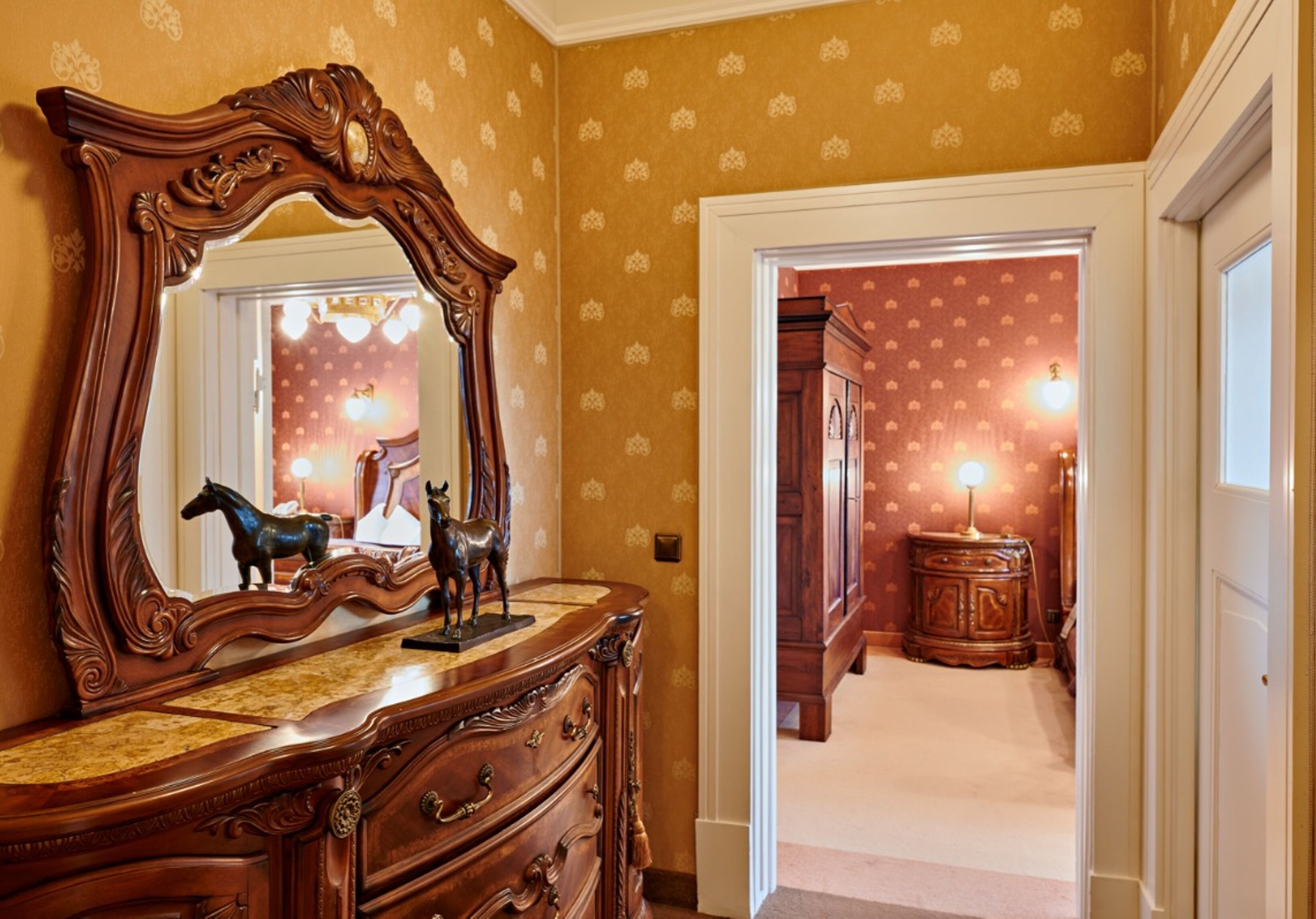 A room with a large ornate mirror, a wooden dresser, and a door leading to another room with patterned wallpaper and a lamp.
