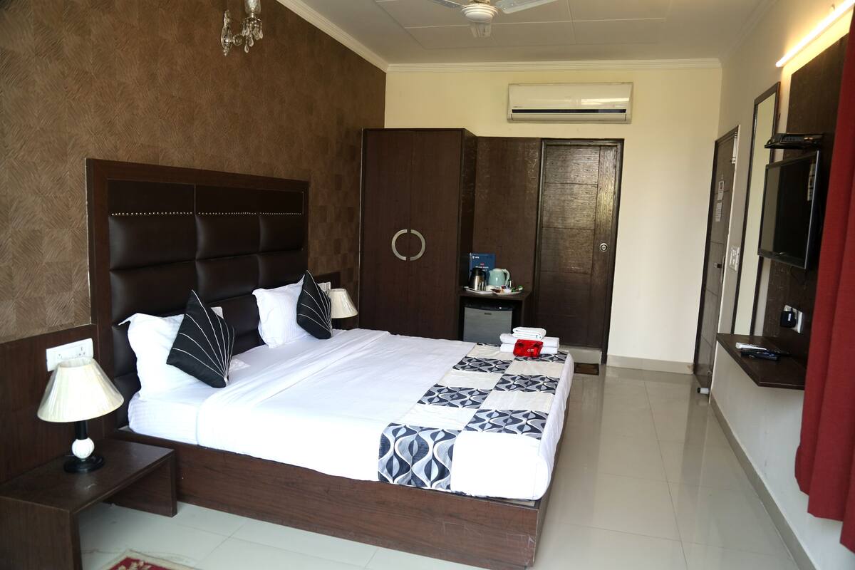 Une chambre d’hôtel avec un grand lit, des tables de chevet, une petite table, un appareil de climatisation, un miroir et un rideau rouge.
