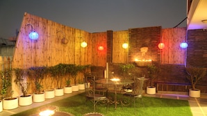 A wooden fence with potted plants, a glass table with chairs, and a small fire feature. - Anantkoti Hotel (New Delhi)