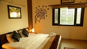 A neatly made bed with a white and brown bedspread, a wall-mounted tree decal with birds, and a window with shutters. - Anantkoti Hotel (New Delhi)