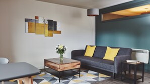 Un salon moderne avec un canapé gris, des coussins jaunes, une table basse en bois et des œuvres d’art abstraites accrochées au mur.