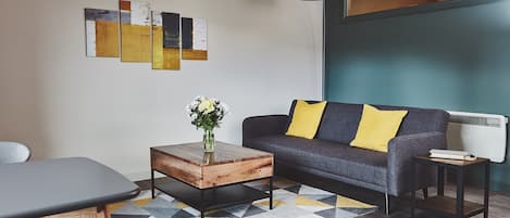 Ein modernes Wohnzimmer mit grauem Sofa, gelben Kissen, einem Holztisch und abstrakter Wandkunst.