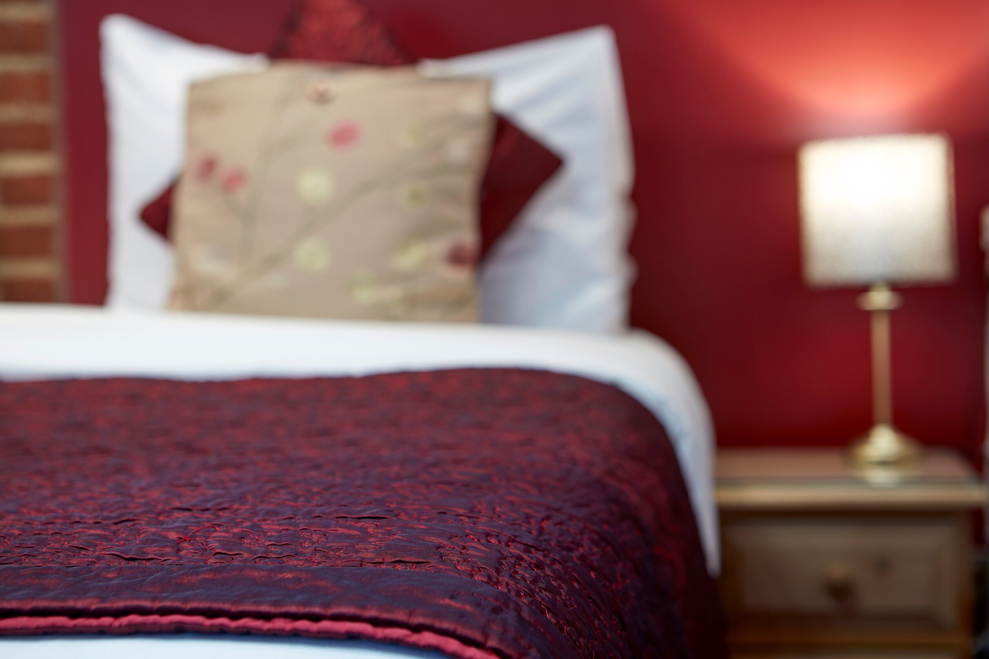 Een bed met een rode deken, een kussen met een bloemenprint en een bedlampje op een houten nachtkastje.