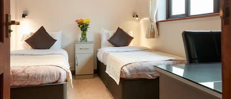Une chambre avec deux lits, un bureau et une chaise. On y trouve une table de chevet sur laquelle est posé un vase de fleurs.
