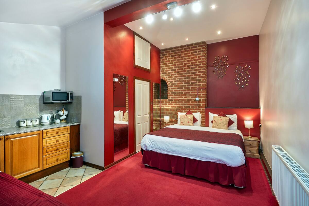 Ein modernes Hotelzimmer mit rotem und weißem Farbkonzept, einer Wand aus Ziegelsteinen, einem großen Bett mit roter Tagesdecke, einer Holzkommode, einer Mikrowelle und einer kleinen Küchenzeile.