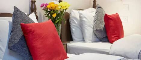 A hotel room with two beds, each with white bedding and multiple pillows in red, grey, and white. A vase with yellow flowers is placed between the beds.