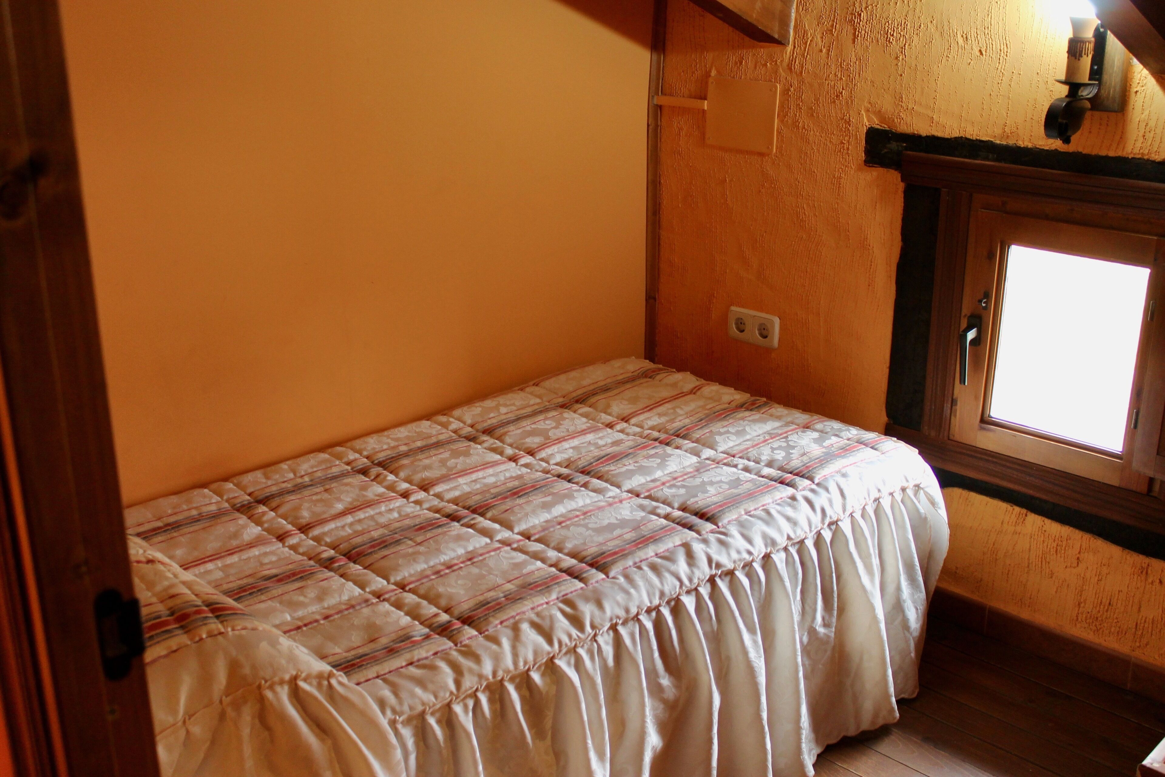 A small, single-bed room with a wooden headboard, a window with a view, and a wall-mounted light fixture.