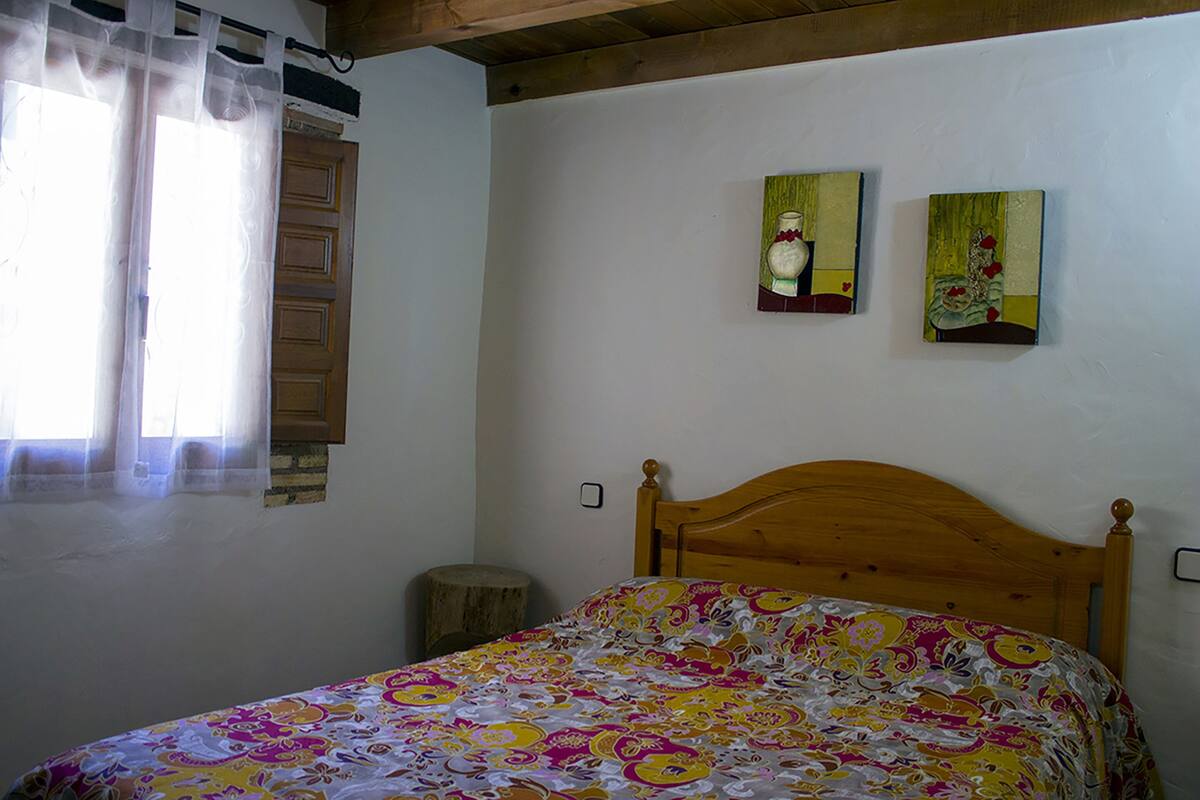 A bedroom with a wooden bed, a window with sheer curtains, and two paintings on the wall.