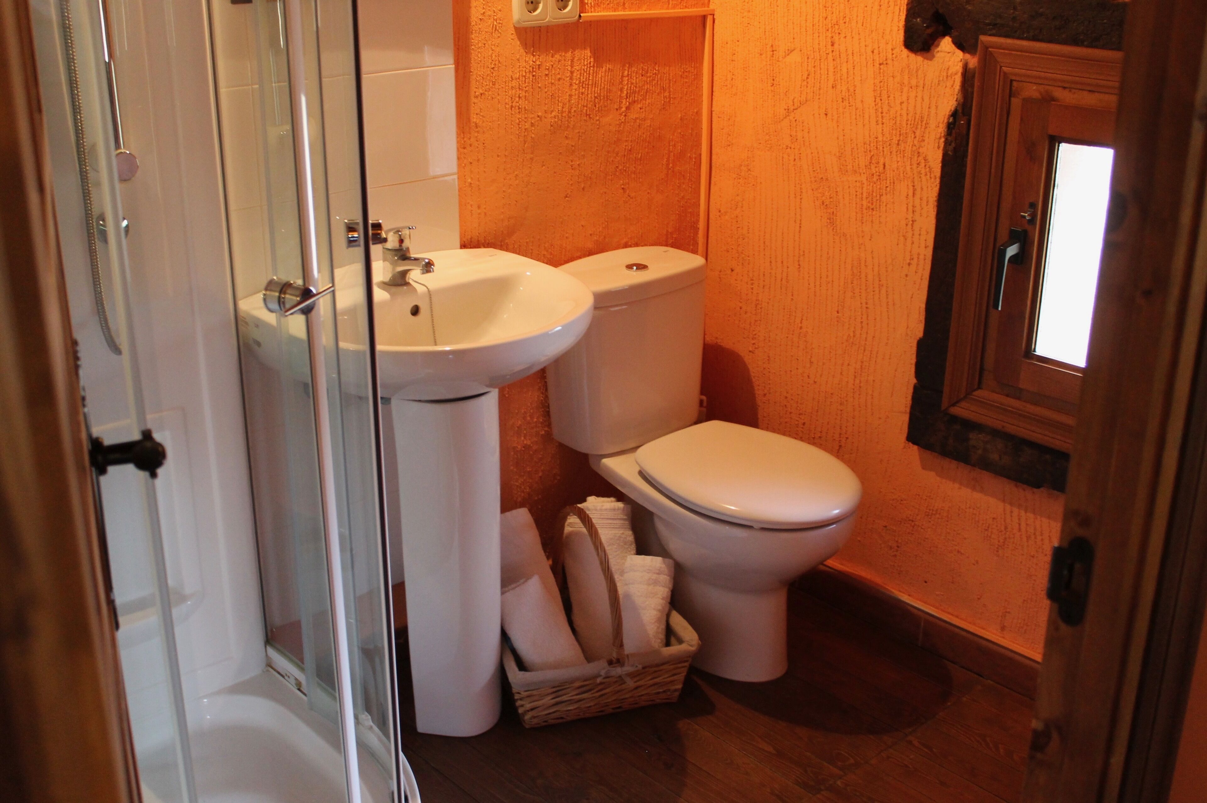 A bathroom with a glass-enclosed shower, two sinks, a toilet, and a wooden door.