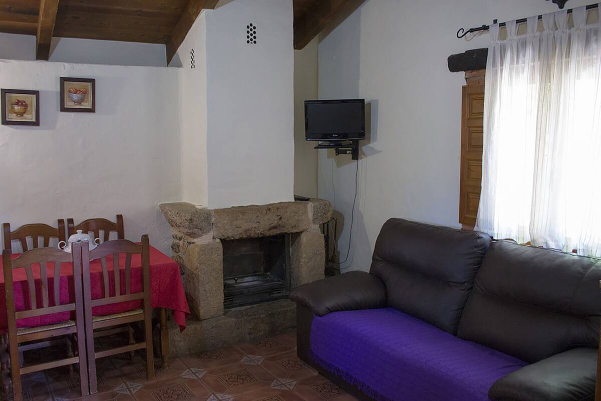 A cozy living room with a stone fireplace, a leather sofa, a dining table with chairs, and a television mounted on the wall.