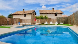 Une piscine dotée d’une terrasse carrelée, entourée d’une clôture en bois et bordée de deux maisons aux toits couverts de tuiles.