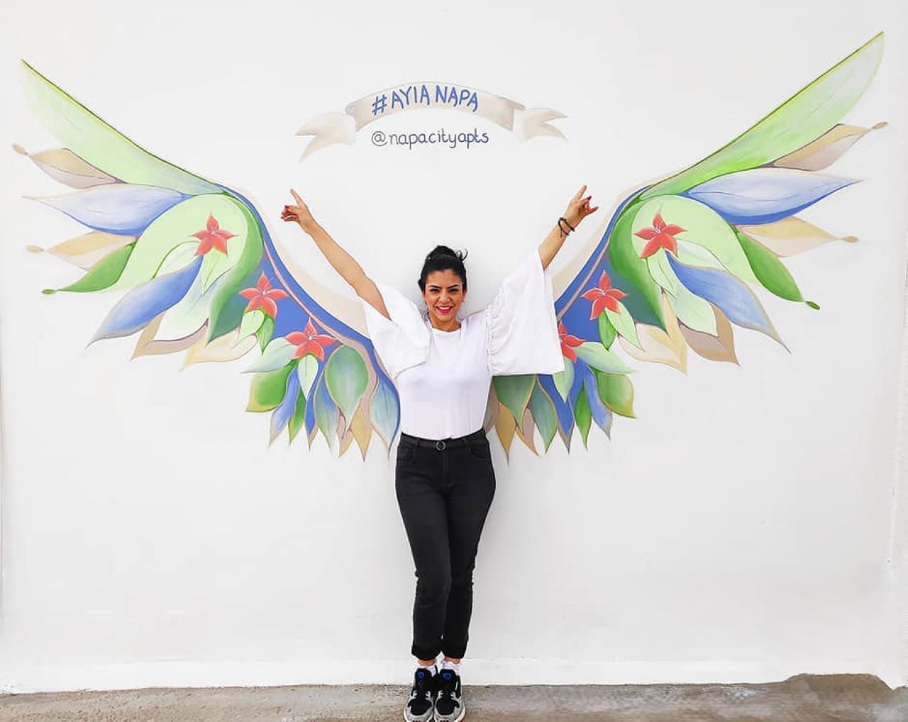 Une personne se tient devant un mur orné d’une fresque colorée représentant des ailes, et une banderole avec du texte est accrochée au mur.